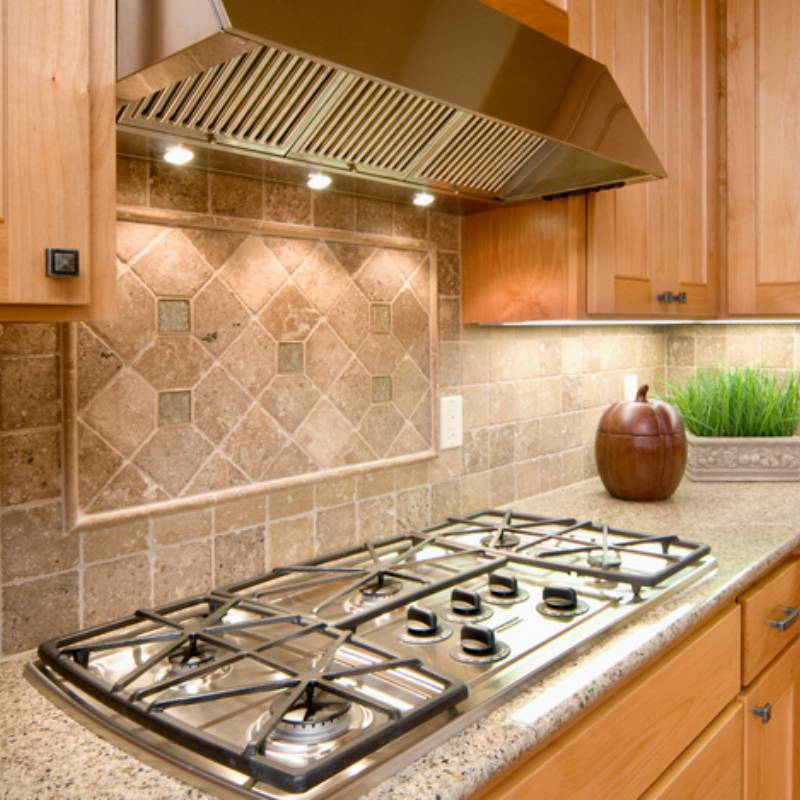 Modern kitchen stovetop with stainless steel hood and tile backsplash.